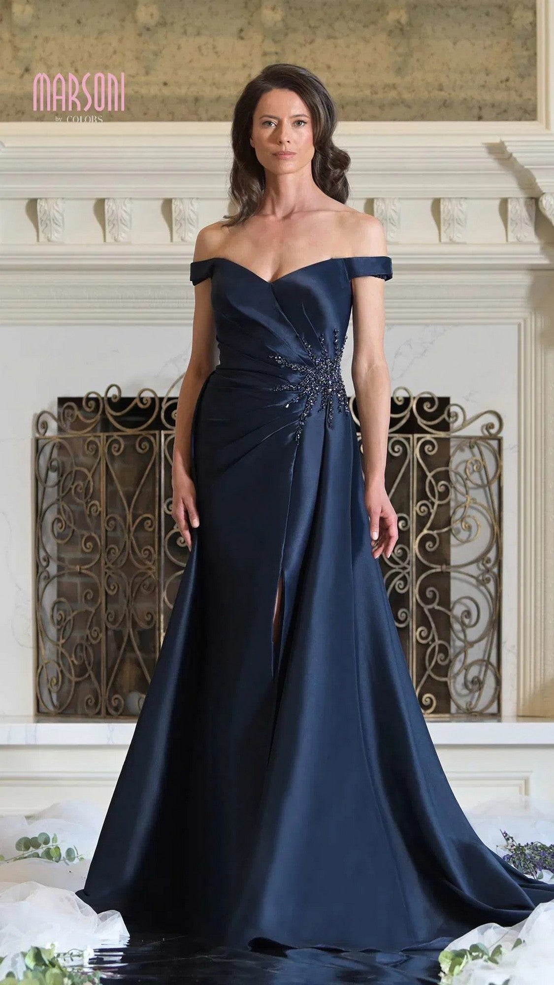 A woman stands indoors in the Marsoni MV1310 long formal dress, a navy off-the-shoulder gown with a mermaid silhouette, front slit, and embellishments, posed before an ornate fireplace.